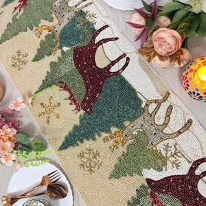 May include: Beaded Christmas table runner with a festive design. Features reindeer, evergreen trees, and snowflakes in shades of green, red, gold, and cream. Decorative table setting with plates, bowls, and utensils.