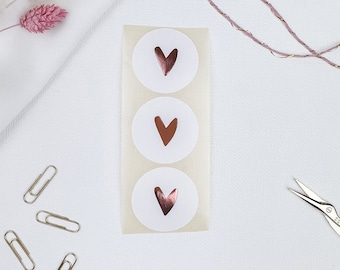 May include: Three white round stickers with a rose gold heart design. The stickers are arranged vertically on a white background.