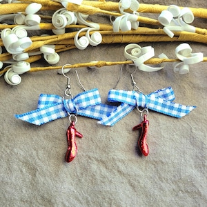 May include: A pair of dangle earrings featuring blue and white gingham bows and small red high-heeled shoe charms. The earrings hang from silver-colored hooks, with a whimsical and playful design.