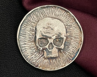 May include: A silver coin with a detailed skull design. The skull is centered on the coin and surrounded by a textured, radiating pattern.