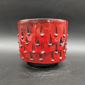 Könnte beinhalten: Ein roter Keramiktopf mit schwarzer Glasur, der ein sich wiederholendes Muster aus kleinen, spitzen Formen aufweist. Der Topf hat einen abgerundeten Boden und einen leicht ausgestellten Rand.
