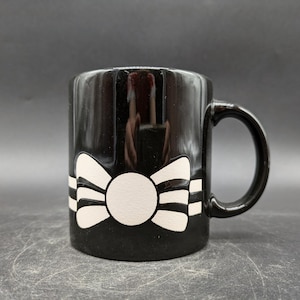 Könnte beinhalten: Schwarze Keramiktasse mit einem weißen Schleifendesign. Die Tasse hat einen Henkel und ist für heiße Getränke geeignet.
