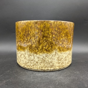 Könnte beinhalten: Zylindrischer Keramiktopf mit einer gesprenkelten, strukturierten Oberfläche. Die obere Hälfte ist dunkelbraun, geht unten in ein helleres Beige über. Der Topf hat eine weite Öffnung und einen abgerundeten Boden.