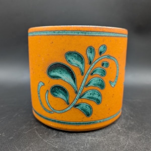 Könnte beinhalten: Orangefarbene Keramiktasse mit türkisem floralem Design. Die Tasse hat eine zylindrische Form mit einem türkisfarbenen Band oben und unten. Das florale Design ist eine türkisfarbene Ranke mit Blattformen.