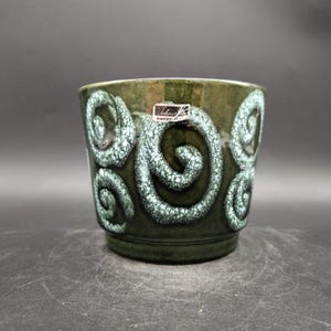 Pode incluir: Vaso de cerâmica verde escuro com um padrão de redemoinho texturizado em azul claro. O vaso tem formato cilíndrico e uma pequena etiqueta. Uma peça decorativa para plantas de interior, com cerca de 15 cm de altura.