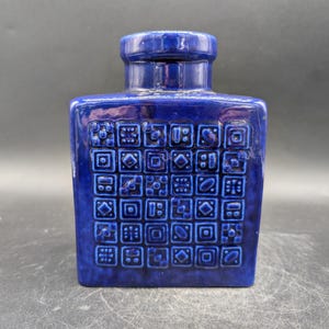 Könnte beinhalten: Eine quadratische, kobaltblaue Keramikvase mit einem strukturierten, geometrischen Muster. Die Vase hat eine rechteckige Form mit kurzem, zylindrischem Hals und flacher Oberseite. Die Oberfläche ist glänzend, und das Design besteht aus kleinen, sich wiederholenden Quadraten.