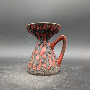 May include: A ceramic vase with a handle, featuring a textured, volcanic-like design. The vase is primarily dark brown with red and black accents. The handle is red, and the opening is wide.