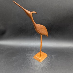 May include: A wooden bird sculpture with a long, slender beak and a curved neck. The bird is perched on a square wooden base. The sculpture is made of a warm brown wood and has a smooth finish. The bird's eye is a small, round detail.