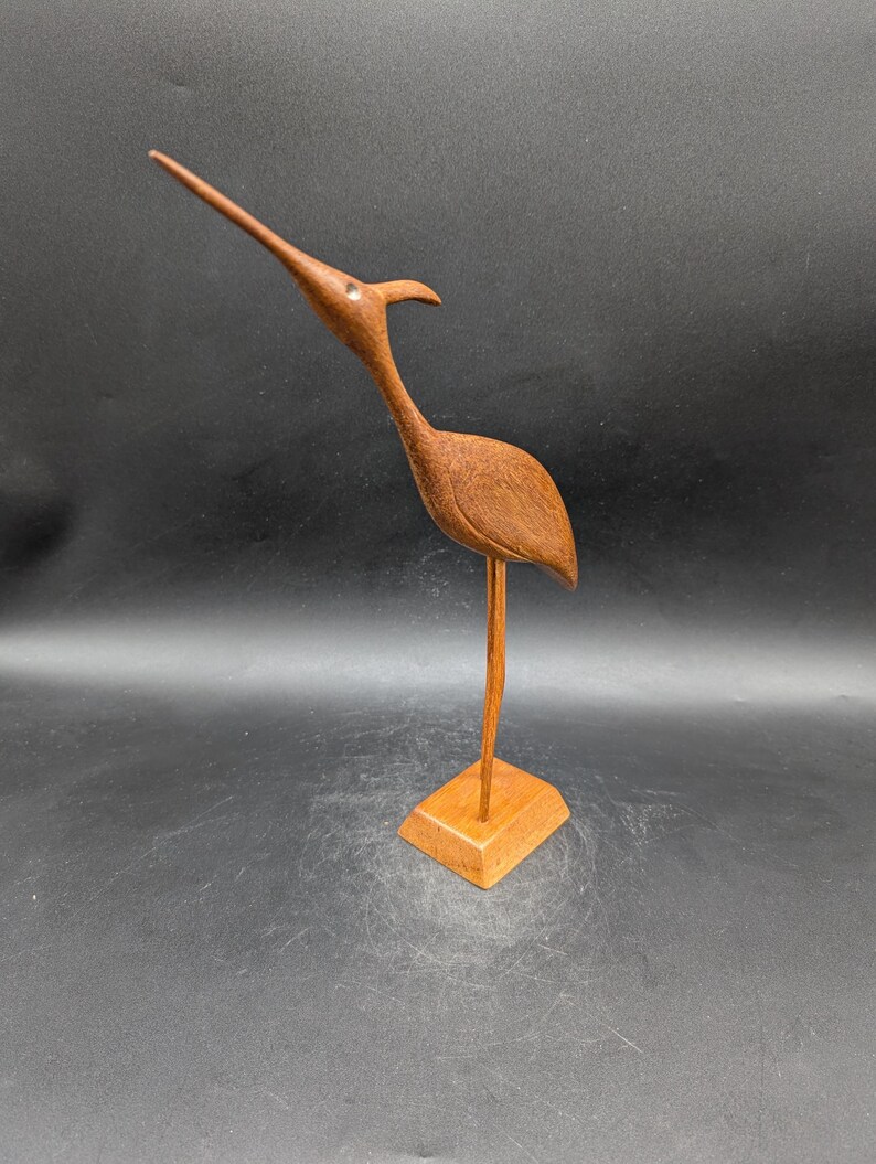 May include: A wooden bird sculpture with a long, slender beak and a curved neck. The bird is perched on a square wooden base. The sculpture is made of a warm brown wood and has a smooth finish. The bird's eye is a small, round detail.