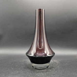 May include: A tall, elegant vase with a flared base and a slender neck. The vase features a gradient of colors, transitioning from a dark, opaque base to a translucent, reddish-purple upper portion. The vase is made of glass.