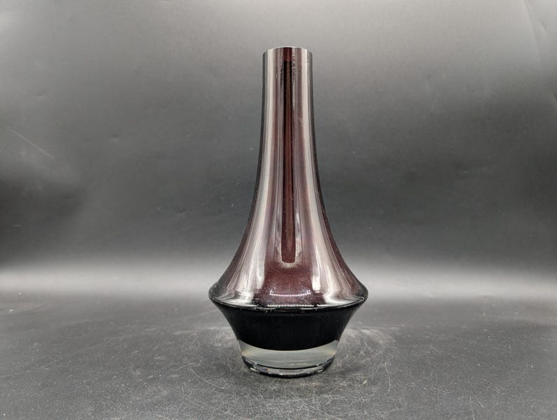 May include: A tall, elegant vase with a flared base and a slender neck. The vase features a gradient of colors, transitioning from a dark, opaque base to a translucent, reddish-purple upper portion. The vase is made of glass.