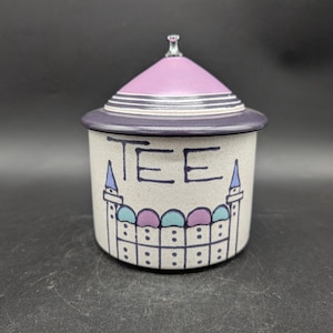 Könnte beinhalten: Eine Keramik-Teedose mit Deckel. Die Dose ist cremefarben mit dem Wort "TEE" in schwarzen Buchstaben. Der Deckel ist lavendelfarben mit einem silbernen Band. Die Dose hat ein dekoratives Burgdesign in Blau, Türkis und Lila.