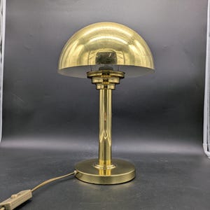 Könnte beinhalten: Eine goldfarbene Tischlampe mit einem kuppelförmigen Schirm. Die Lampe hat ein schlichtes, elegantes Design und eine klassische Optik.