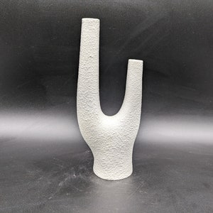 May include: White ceramic vase with a unique, abstract design. The vase has a textured surface and two openings for flowers.