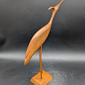 May include: A wooden bird sculpture with a long neck and beak, standing on a square base. The bird is a warm brown color, and the sculpture has a smooth finish. The bird's head is tilted upwards, giving it a graceful appearance. The sculpture is a decorative item.