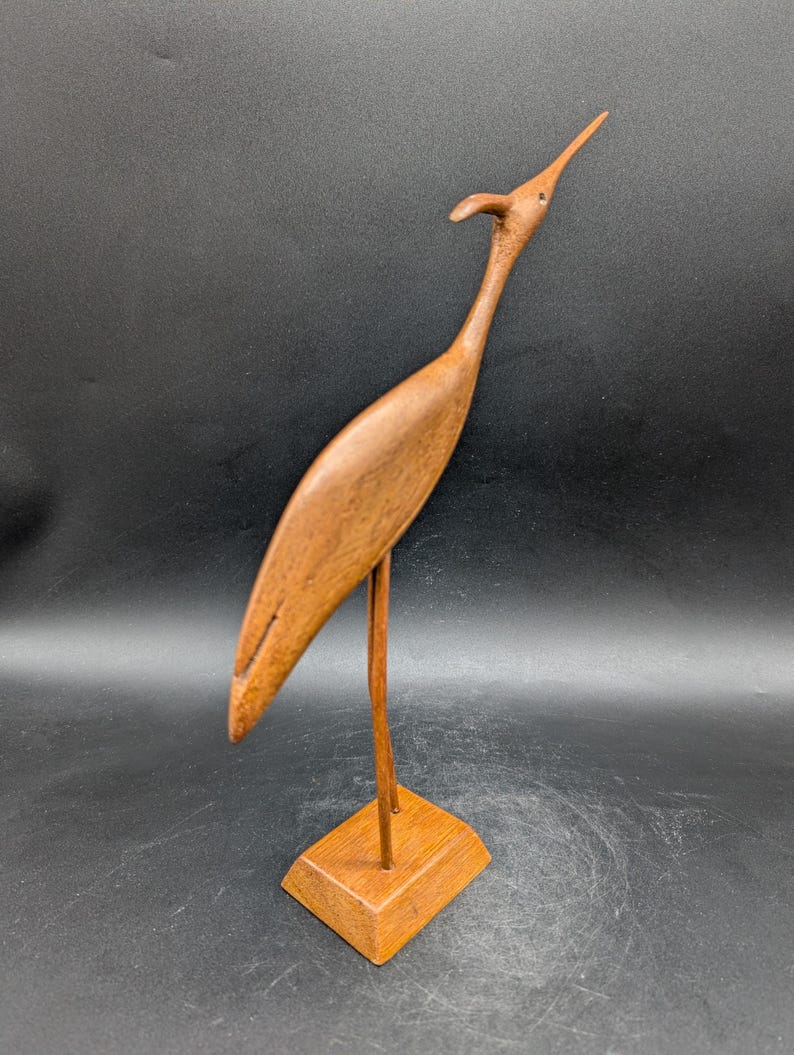 May include: A wooden bird sculpture with a long neck and beak, standing on a square base. The bird is a warm brown color, and the sculpture has a smooth finish. The bird's head is tilted upwards, giving it a graceful appearance. The sculpture is a decorative item.