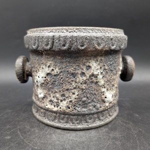 Puede incluir: Una maceta cilíndrica de cerámica con una superficie texturizada y moteada. Tiene dos asas pequeñas y redondeadas y bandas decorativas en la parte superior e inferior. La paleta de colores incluye tonos marrones, negros y blancos. Aproximadamente 10 cm de altura.