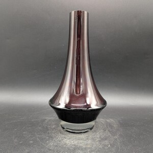 May include: A tall, elegant vase with a flared shape. The vase features a gradient of colors, transitioning from a deep purple at the base to a lighter, almost iridescent hue towards the top. The base is black, and the bottom is clear glass.