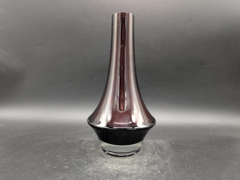 May include: A tall, elegant vase with a flared shape. The vase features a gradient of colors, transitioning from a deep purple at the base to a lighter, almost iridescent hue towards the top. The base is black, and the bottom is clear glass.
