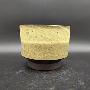 May include: A ceramic planter with a speckled, light brown glaze. The planter has a wide, round opening and a slightly flared base. The base is a darker brown color.