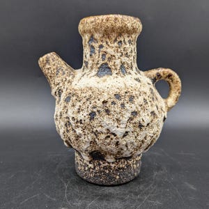 Könnte beinhalten: Eine kleine, braune und weiße Keramikvase mit einem gesprenkelten Überzug. Die Vase hat einen runden Körper und einen Ausguss auf der einen Seite. Sie hat einen Griff auf der anderen Seite.