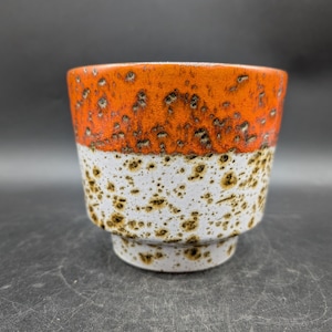 Könnte beinhalten: Eine Keramiktasse mit gesprenkeltem Design. Das obere Drittel der Tasse ist orange, der mittlere Abschnitt ist weiß mit braunen Sprenkeln und der Boden ist weiß. Die Tasse hat eine zylindrische Form mit einem kleinen Sockel.