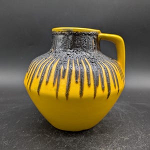 May include: A yellow ceramic vase with a black glaze design. The vase has a wide mouth and a handle.