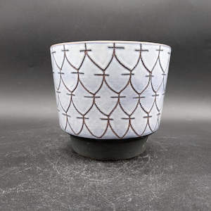 Könnte beinhalten: Ein Keramik-Pflanztopf mit einem hellblauen und braunen geometrischen Muster. Der Topf hat eine breite, zylindrische Form mit einem dunkelgrauen Sockel. Das Muster ist ein sich wiederholendes Design aus miteinander verbundenen Formen. Der Rand ist dunkelbraun.