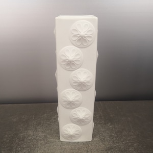 May include: White ceramic vase with a square shape and embossed floral designs.