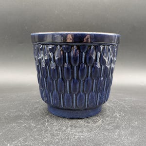 Puede incluir: Un macetero de cerámica azul oscuro con un diseño texturizado de pequeños óvalos alargados. El macetero tiene una forma simple y cilíndrica.