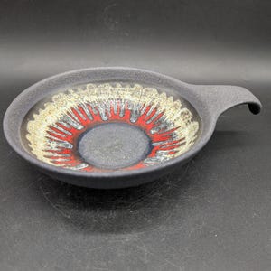 Puede incluir: Un cuenco de cerámica gris oscuro con asa. El interior presenta un diseño circular con detalles en rojo, blanco y amarillo. El cuenco tiene un acabado mate y un borde ligeramente elevado, adecuado para servir o exhibir.