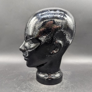 May include: A black, glossy mannequin head with a smooth, reflective surface. The head is a side profile, showing the nose, ear, and jawline. The base is a solid, rounded shape.