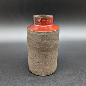 May include: A small, dark brown ceramic vase with a red rim. The vase has a textured surface and a rounded shape.