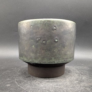 Könnte beinhalten: Ein zylindrischer Keramik-Pflanztopf mit dunkler, gesprenkelter grün-grauer Oberfläche. Der Pflanztopf hat eine breite, abgerundete Oberseite und steht auf einem kleineren, dunkelbraunen Sockel. Die Oberfläche hat ein strukturiertes Aussehen mit kleinen, unregelmäßigen Unebenheiten.
