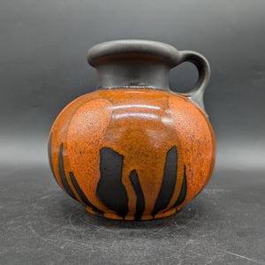 Könnte beinhalten: Eine Keramikvase mit rundem Körper und Henkel. Die Vase ist hauptsächlich orange mit schwarzen, organisch geformten Mustern. Der Rand und der Henkel sind dunkelgrau. Die Vase ist etwa 18 cm hoch.