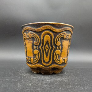 May include: A brown ceramic planter with a decorative design of stylized swirls and diamonds. The planter has a scalloped rim.
