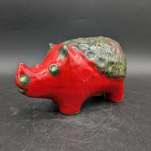 Könnte beinhalten: Ein rotes Keramik-Sparschwein mit grüner und schwarzer Glasur. Das Sparschwein ist in Form eines Schweins mit einem eingerollten Schwanz und einer Schnauze.
