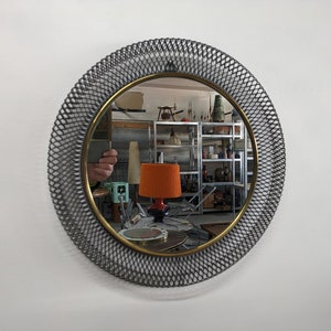 Puede incluir: Un espejo redondo con un marco de malla de alambre negro y un borde dorado. El espejo refleja una habitación con estantes y varios objetos.
