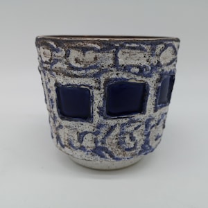 Puede incluir: Un macetero de cerámica blanca con una superficie texturizada y cuadrados esmaltados azules. El macetero tiene un aspecto rústico y hecho a mano.