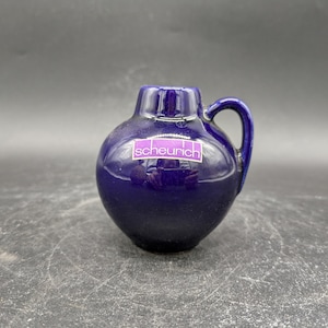 Könnte beinhalten: Ein kleiner, dunkelblauer Keramik Krug mit Henkel. Der Krug hat ein violettes Etikett mit der Aufschrift "scheurich".