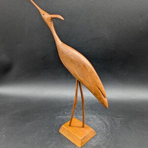 May include: A wooden bird sculpture with a long neck and beak, standing on a small square base. The bird is a warm brown color, with a smooth finish. The sculpture is a decorative item.