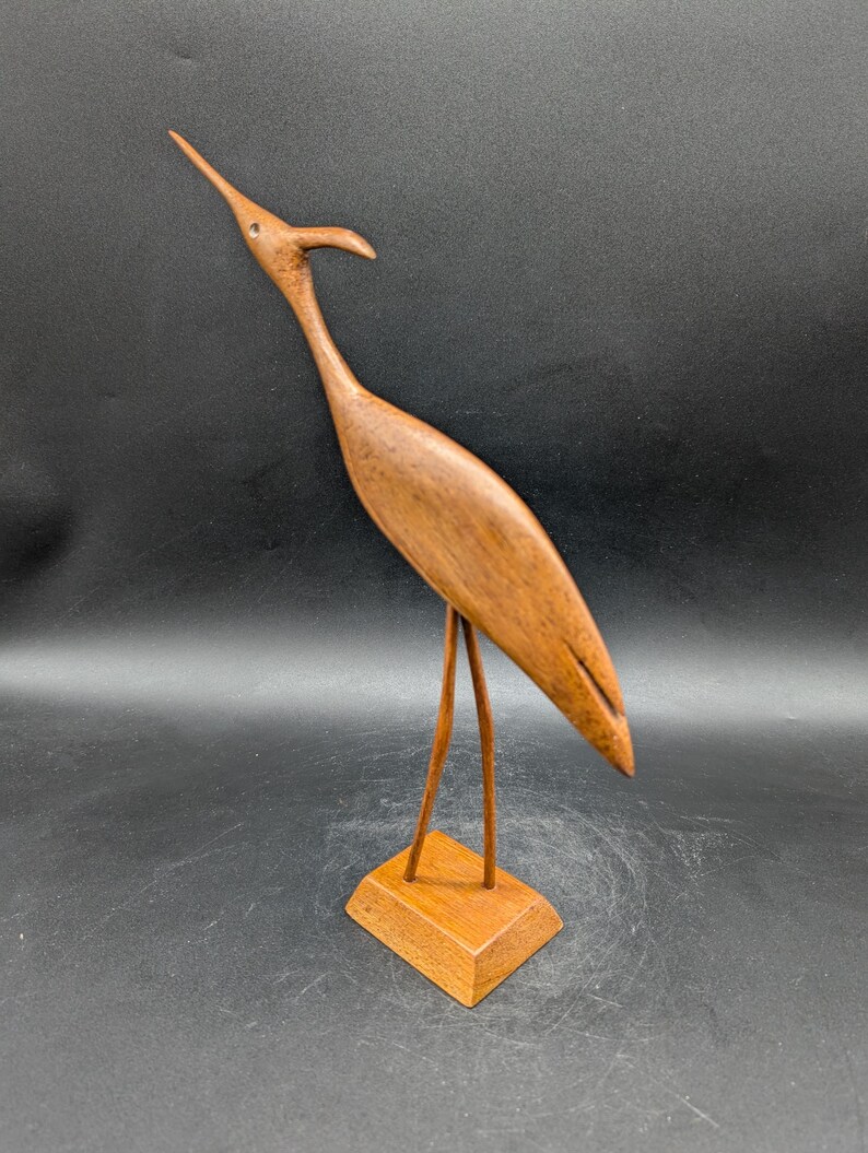 May include: A wooden bird sculpture with a long neck and beak, standing on a small square base. The bird is a warm brown color, with a smooth finish. The sculpture is a decorative item.