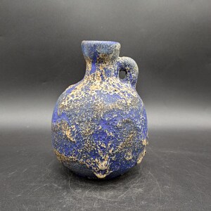 May include: A small, blue ceramic vase with a textured surface and a handle. The vase has a round body and a narrow neck.