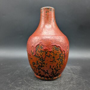 May include: A small, red ceramic vase with a textured surface and a narrow neck. The vase has dark, mottled patterns near the base and neck, adding visual interest. The vase is likely for decorative purposes.