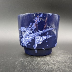 May include: A blue ceramic planter with white splatter paint design. The planter has a rounded bottom and a slightly flared top.