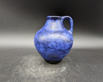 May include: A small blue ceramic pitcher with a speckled glaze and a handle. The pitcher is a light blue color with dark blue speckles throughout.