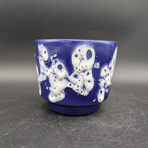 Könnte beinhalten: Ein blauer Keramiktopf mit weißen abstrakten Mustern. Der Topf hat eine abgerundete Form und eine glatte, glänzende Oberfläche.