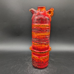 Puede incluir: Una jarra de cerámica roja y naranja con una superficie texturizada y un asa. La jarra tiene un diseño único con un aspecto en capas.