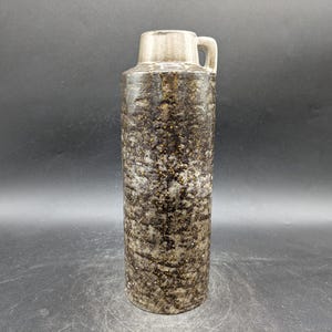 May include: A tall, cylindrical ceramic vase with a textured, speckled surface in shades of brown, gray, and gold. The vase has a light-colored top with a small handle. The design is reminiscent of mid-century modern pottery.