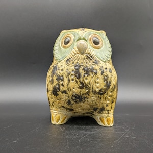 May include: A ceramic owl figurine with brown, black and green glaze. The owl has large, round eyes with black pupils and a beak.